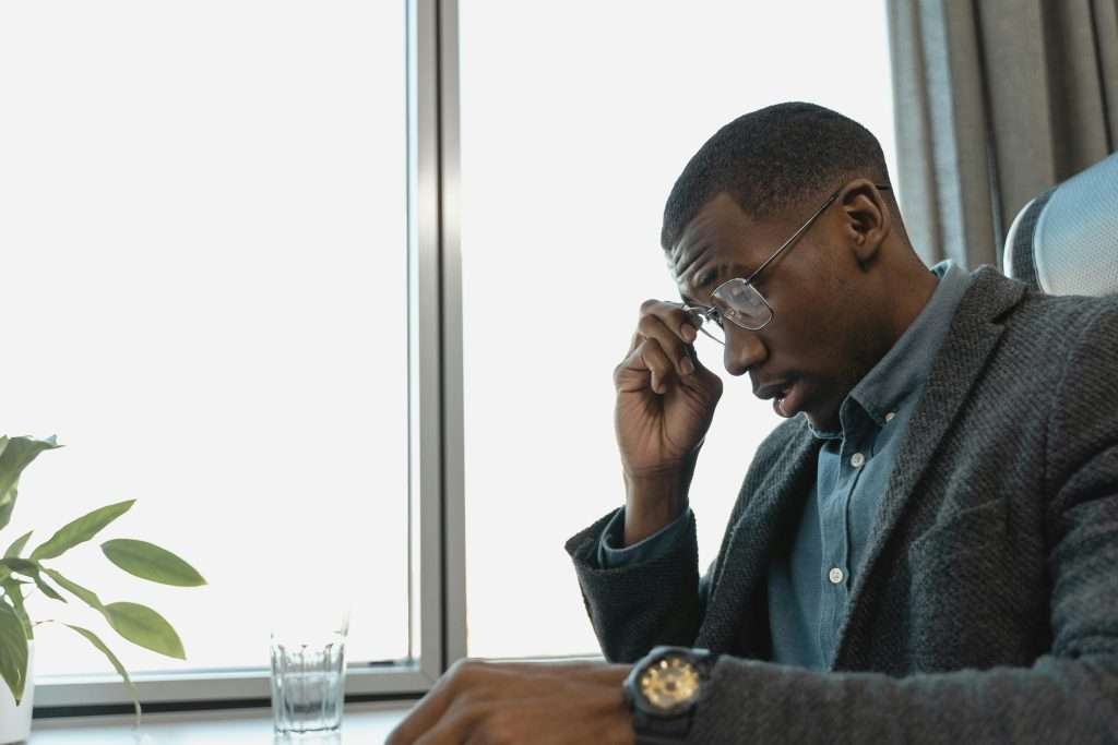 Exhausted businessman in glasses contemplating at office desk near window.